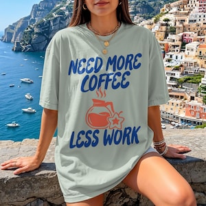 Puede incluir: Camiseta verde claro con el texto azul "NEED MORE COFFEE LESS WORK". El diseño incluye una cafetera y una estrella naranja. La camiseta es de corte holgado y relajado.