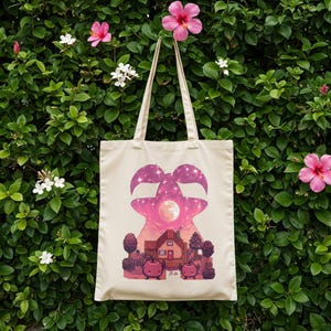 May include: Cream-coloured tote bag featuring a pink and purple graphic. The graphic depicts a stylized shape with a starry sky design, a full moon, and a pixelated house with trees and two red apple characters. The bag has long handles.