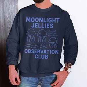 以下が含まれることがあります： ライトブルーの「MOONLIGHT JELLIES OBSERVATION CLUB」の文字が入ったネイビーブルーのスウェットシャツ。 デザインにはクラゲのイラストと「Annual meet up, 8pm sharp」の文字が含まれています。