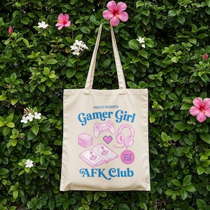 以下が含まれることがあります： ベージュ色のトートバッグ。テキスト「Proud Member Gamer Girl AFK Club」が青とピンクでプリントされています。ゲームコントローラー、ヘッドフォン、ハート、本のイラストがデザインされています。バッグにはベージュ色のハンドルが2つ付いています。