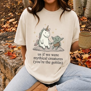Può includere: Una t-shirt color crema con la grafica di un unicorno bianco e un goblin verde seduti insieme su una roccia. Il testo sotto l'illustrazione recita "us if we were mythical creatures (you're the goblin)" in un semplice carattere nero.