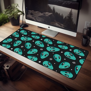 May include: Black desk mat featuring a repeating pattern of teal alien faces and white stars. The desk mat is rectangular and designed to protect a desk surface. The alien faces have large black eyes and are smiling.
