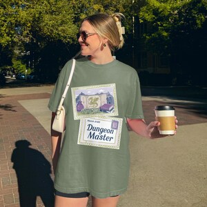 Op de afbeelding: Olijfgroen t-shirt met twee grafische ontwerpen. Het bovenste ontwerp toont een kaart, een draak en boeken. Het onderste ontwerp luidt "Dungeon Master" met "Maart 2020" erboven. Een koffiekopje wordt in de hand gehouden.