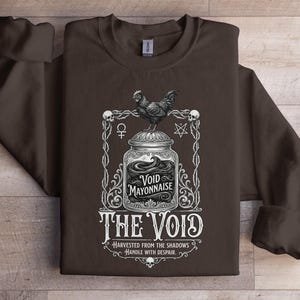 May include: A dark brown sweatshirt featuring a graphic design. The design includes a jar labeled "Void Mayonnaise" with a rooster on top, framed by decorative elements and the text "The Void". The text "Harvested from the Shadows, Handle with Despair" is also included.