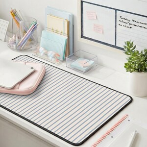 May include: A desk setup featuring a striped desk mat in cream and blue, a pink tablet case, and clear organizers holding pens, notebooks, and sticky notes. A whiteboard with notes and a small potted plant complete the scene.