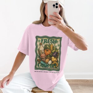 Könnte beinhalten: Hellrosa T-Shirt mit einem Aufdruck mit der Aufschrift "FRESH FARM LIFE" und einem Korb mit Produkten. Der Korb enthält eine Milchflasche, einen Kürbis und anderes Gemüse. Das T-Shirt hat auch Text unten.