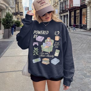 May include: Navy blue sweatshirt with the text "POWERED BY" and illustrations of gaming consoles, a cozy gamer, and the words "Tea and Mild Anxiety." A beige cap with text is worn.