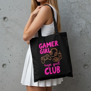 以下が含まれることがあります： ピンクとオレンジ色の「GAMER GIRL rage quit CLUB」という文字が入った黒いトートバッグ。ゲーム用ヘッドセット、コントローラー、食べ物のイラストが描かれています。バッグは人が持っています。