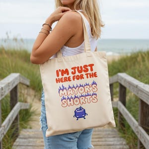 May include: Cream-colored tote bag with the text "I'M JUST HERE FOR THE MAYOR'S SHORTS" in red and blue. The bag features a pixelated blue character. The bag is carried by a person on a wooden walkway near the beach.