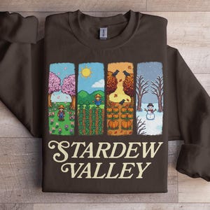 May include: A brown sweatshirt featuring a pixelated design of four seasonal scenes. The scenes depict spring, summer, autumn, and winter, with the text "Stardew Valley" in a stylized font at the bottom.