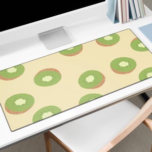 May include: A rectangular desk mat featuring a repeating pattern of green and brown kiwi fruit slices on a light yellow background. The mat is placed on a white desk, with a computer monitor, pencils, and other office supplies visible.