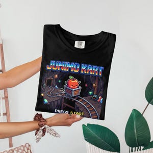 May include: Black t-shirt featuring a pixelated graphic of a mine cart with an apple character, the text "JUNIMO KART", and "PRESS START". The design is in a retro video game style.