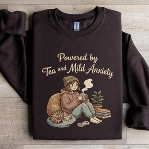 May include: A dark brown sweatshirt featuring the phrase "Powered by Tea and Mild Anxiety." The illustration depicts a person, a cat, tea, and books. The sweatshirt has a crew neck and long sleeves.