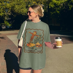 May include: Sage green t-shirt featuring a fall-themed graphic. The graphic includes a scarecrow, pumpkins, corn stalks, and the text "IT'S A GOOD LIFE... I GUESS" within a brown border. The shirt is a relaxed fit.