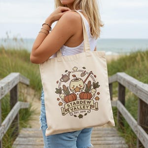 May include: Cream-colored tote bag featuring a circular graphic with pumpkins, corn, carrots, and a watering can. The graphic includes the text "STARDEW VALLEY EST. 2016" on a banner. The bag is carried over the shoulder.