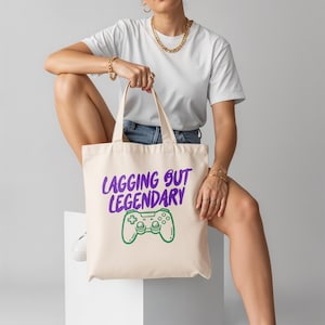 以下が含まれることがあります： 紫色の「LAGGING BUT LEGENDARY」の文字と緑色のゲームコントローラーのグラフィックがプリントされた、クリーム色のトートバッグ。バッグには2つのハンドルがあり、様々なアイテムを持ち運ぶのに適した丈夫な生地で作られています。