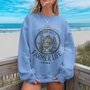 Könnte beinhalten: Hellblaues Sweatshirt mit einem kreisförmigen Aufdruck "Stardew Valley Farmer Life Since 2016". Die Grafik zeigt eine Farm mit Scheune, Bäumen und Gemüse. Das Sweatshirt hat einen Rundhalsausschnitt und lange Ärmel.