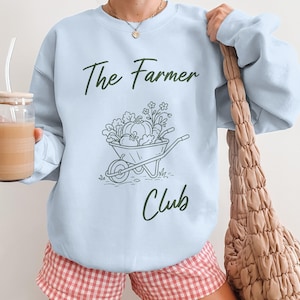 Könnte beinhalten: Hellblaues Sweatshirt mit dem Text "The Farmer Club" in dunkelgrüner Schrift. Eine Grafik einer Schubkarre mit Gemüse und Blumen befindet sich unter dem Text. Die Person trägt rot-weiß karierte Shorts.