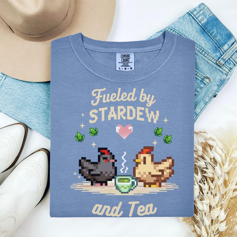a blue t-shirt with a pixel art design of two chickens and a cup of tea, along with the text "Fuelled by Stardew and Tea".