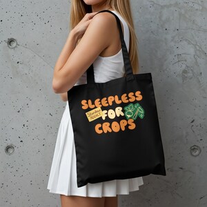 May include: Black tote bag with the text "SLEEPLESS FOR CROPS" in orange and a small graphic. The bag also features a yellow sticky note that says "Stardew Addict". The bag is carried over the shoulder.