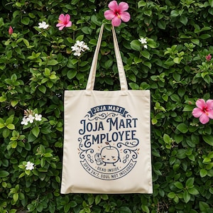 May include: A cream-colored tote bag with the text "JOJA MART EMPLOYEE" in navy blue, with an illustration of a sad apple. The bag has long handles and is set against a backdrop of green foliage and pink flowers.