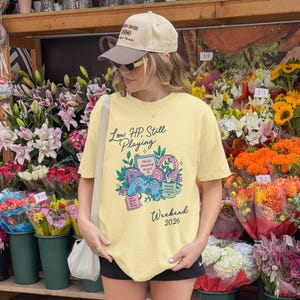 Puede incluir: Una camiseta amarillo claro con el texto "Low HP, Still Playing" y un gráfico de un mando de videojuegos, auriculares y una taza. La camiseta también tiene la palabra "Weekend" y el número "2026".
