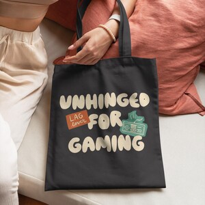 以下が含まれることがあります： 「UNHINGED FOR GAMING」の文字が入った黒いトートバッグ。 「LAG lover」のステッカーとコントローラーのグラフィックが特徴です。 ベージュのトップスと白いパンツを着た人が持っています。