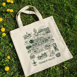May include: Cream-colored tote bag with a green illustration of Pelican Town from Stardew Valley. The design includes a boat, houses, a train, and the text "Pelican Town" and "Stardew Valley". The bag has two cream-colored handles.