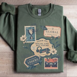 May include: Green sweatshirt with a vintage-style graphic featuring stamps, flowers, and text. The stamps include a trophy, the words "GLOBAL ESPORTS LEAGUE", and "ERROR 404: SOCIAL LIFE NOT FOUND". The sweatshirt has a crew neck and long sleeves.