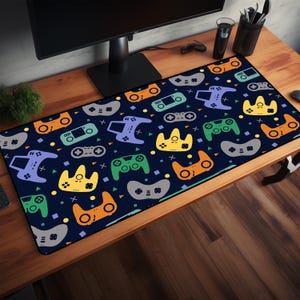May include: A rectangular desk mat with a navy blue background and a pattern of colorful video game controllers. The controllers are in shades of orange, green, purple, yellow, and gray. The mat is on a wooden desk.