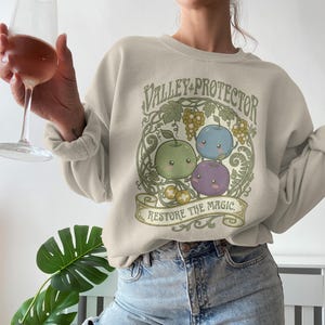 May include: Beige sweatshirt with a circular graphic featuring three cartoon fruits: green apple, blue blueberry, and purple plum. The text "Valley Protector" and "Restore the Magic" are also displayed.