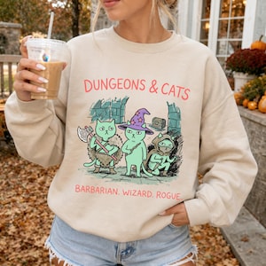 May include: A beige crewneck sweatshirt featuring a graphic print of three light green cats dressed as fantasy characters. The text reads "DUNGEONS & CATS" and "BARBARIAN. WIZARD. ROGUE." The cats are depicted with an axe, a wizard hat, and a dagger.
