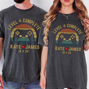 May include: Two black t-shirts with a retro sunset graphic and a video game controller. The text "Level 4 Complete" is above the graphic and "Kate ♥ James 10.4.22" is below the graphic.