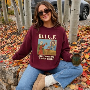 Può includere: Una felpa a maniche lunghe color bordeaux con una stampa grafica di due figure in stile medievale a un tavolo con il testo M.I.L.F. My Inventory Lacks Funds. Il capo è abbinato a jeans azzurri e stivaletti in camoscio color cuoio.