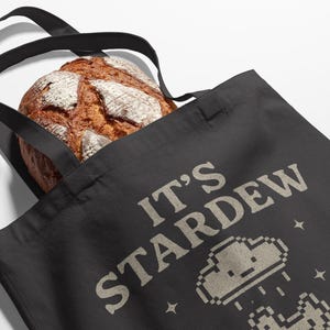 May include: A black tote bag featuring the text "IT'S STARDEW" in beige, accompanied by pixelated cloud and mushroom graphics. A loaf of bread is visible inside the bag.