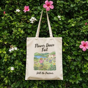 May include: A cream-coloured tote bag with the text "Flower Dance Fail" and "Still No Partner." The bag features a colourful illustration of a person in a field of flowers. The bag has two cream-coloured handles and is hanging against a backdrop of green foliage and flowers.