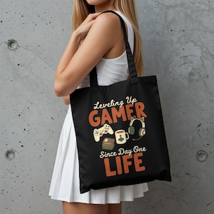 以下が含まれることがあります： オレンジと白で「Leveling Up GAMER Since Day One LIFE」という文字が入った黒いトートバッグ。ゲームコントローラー、ヘッドフォン、コーヒーマグ、本のイラストが描かれています。バッグは人が持っています。