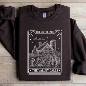 Könnte beinhalten: Dunkelbraunes Sweatshirt mit weißer Grafik einer Farm. Die Grafik zeigt ein Haus, einen Silo und den Text "LIFE ON THE FARM" und "THE VALLEY CALLS". Das Sweatshirt hat einen Rundhalsausschnitt und lange Ärmel.