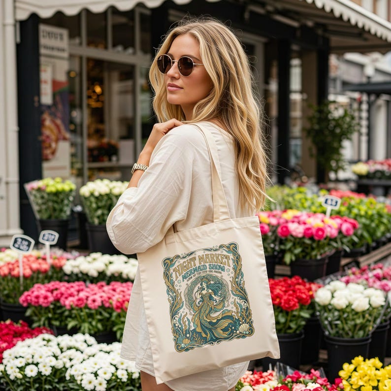 Woman holding tote bag with "Night Market Mermaid Show" graphic near flower stands.