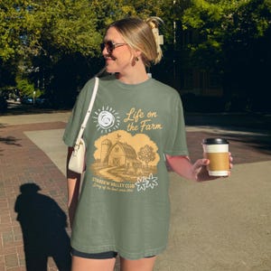 Könnte beinhalten: Olivgrünes T-Shirt mit einem Bauernhof-Motiv. Das Design umfasst eine Scheune, eine Sonne und den Text "Life on the Farm" und "Stardew Valley Club". Das T-Shirt hat eine lockere Passform.
