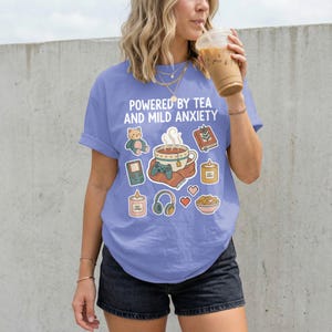 May include: A periwinkle blue t-shirt with the text "POWERED BY TEA AND MILD ANXIETY" and various graphic stickers. The stickers include a teacup, a teddy bear, a book, a video game, candles, headphones, hearts, and a bowl of food.