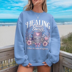 May include: A pale blue sweatshirt featuring the text "Healing Class Energy" in a decorative font. The design includes a computer, headphones, flowers, and the phrase "Where the heals hit different."