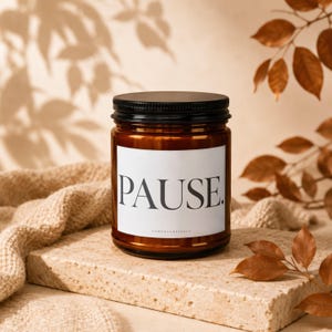 May include: A brown glass candle jar with a black screw-top lid. The white rectangular label on the front features the word PAUSE. in large black serif font, with smaller text below. The jar is cylindrical with a smooth, reflective surface.