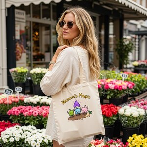 May include: A cream-coloured tote bag featuring the text "Nature's Magic" and a cartoon illustration of three colourful characters. The bag is carried by a person in a white shirt, with a flower shop in the background.