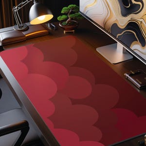 May include: A red desk mat with a repeating cloud-like pattern. The mat is on a wooden desk, with a desk lamp, bonsai tree, and computer monitor in the background. The mat is rectangular.