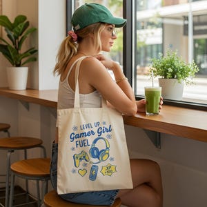 以下が含まれることがあります： クリーム色のトートバッグ。青と黄色の「LEVEL UP Gamer Girl FUEL」の文字。ゲームコントローラー、ヘッドフォン、ハート、バッテリーアイコンのデザイン。緑色の帽子をかぶった人が持っています。