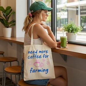 以下が含まれることがあります： 「need more coffee for farming」の文字が青とピンクでプリントされたクリーム色のトートバッグ。太陽、コーヒーカップ、星のグラフィックが特徴です。緑色のキャップをかぶった人がバッグを持っています。