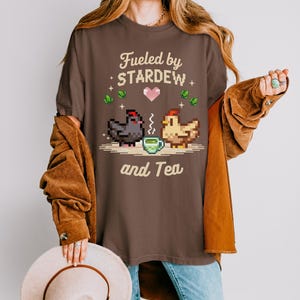 A woman wearing a brown t-shirt with a pixelated design of two chickens and the text "Fuelled by Stardew and Tea" is holding a beige hat.