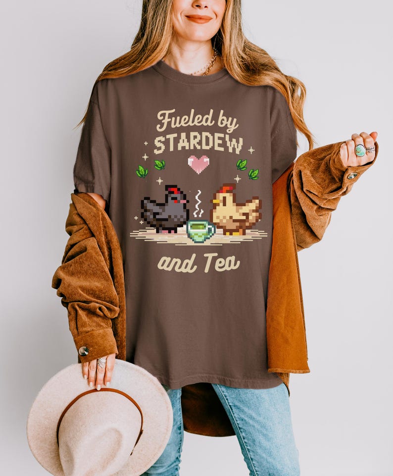 A woman wearing a brown t-shirt with a pixelated design of two chickens and the text "Fuelled by Stardew and Tea" is holding a beige hat.