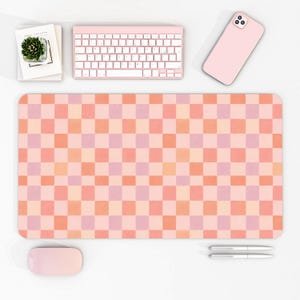 Pode incluir: Tapete de mesa xadrez rosa e laranja com teclado, rato e telefone rosa a condizer. O tapete é retangular e apresenta um padrão de grade de quadrados rosa e laranja. Uma pequena planta suculenta num vaso branco está em cima de livros.